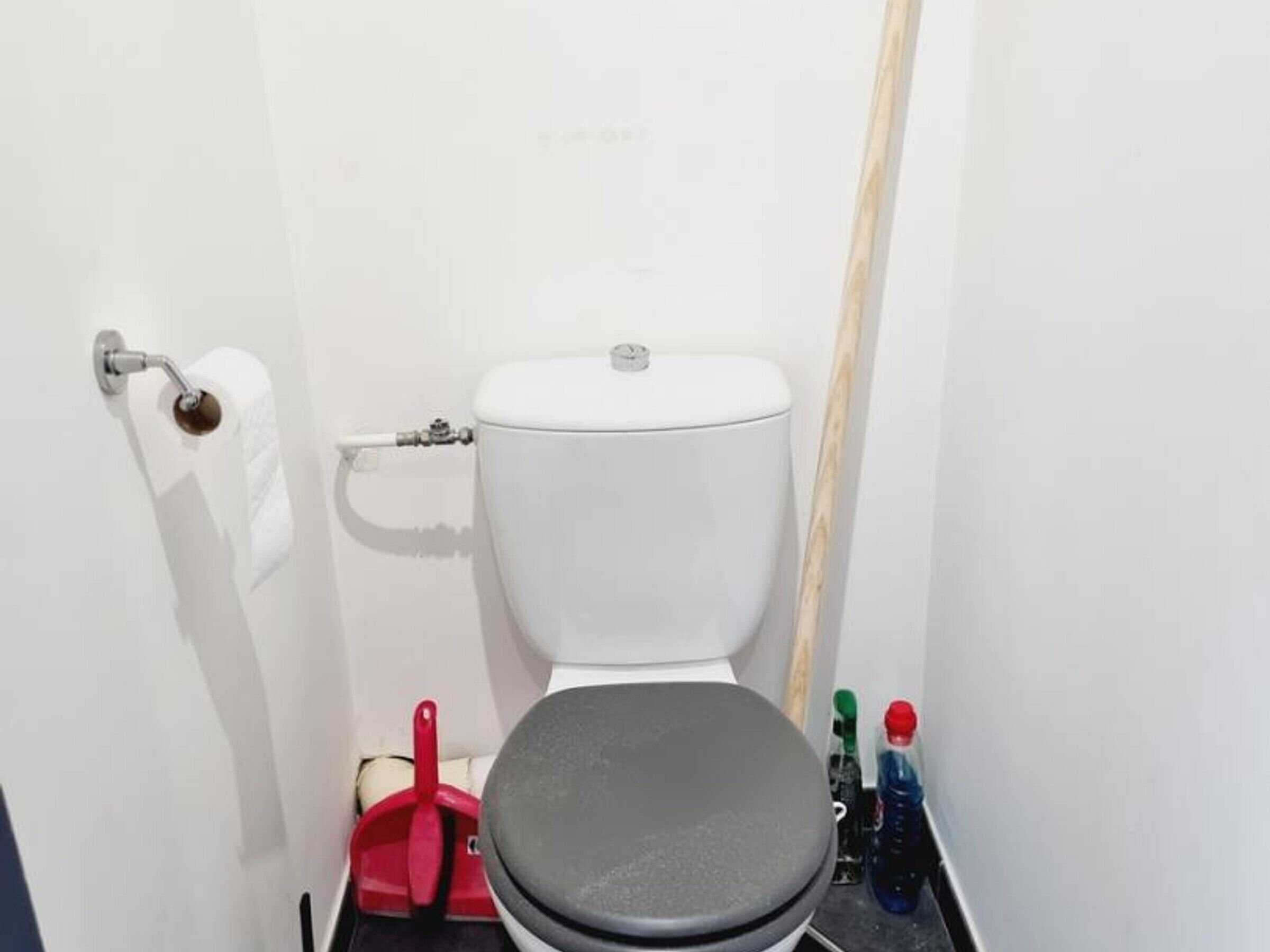 Une petite salle de bain blanche, avec des toilettes, une brosse de nettoyage rouge et une bouteille d’eau posée sur le sol.