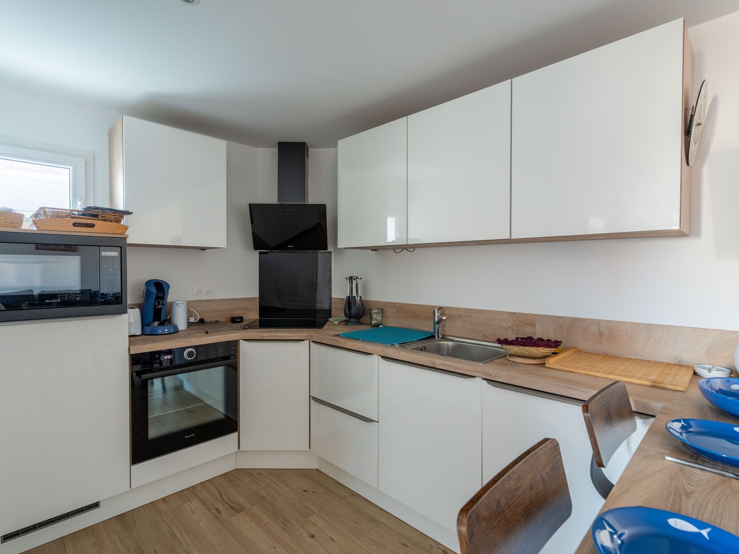 A modern kitchen with white cabinets, a black range hood, a built-in oven, a coffee maker, and a sink.