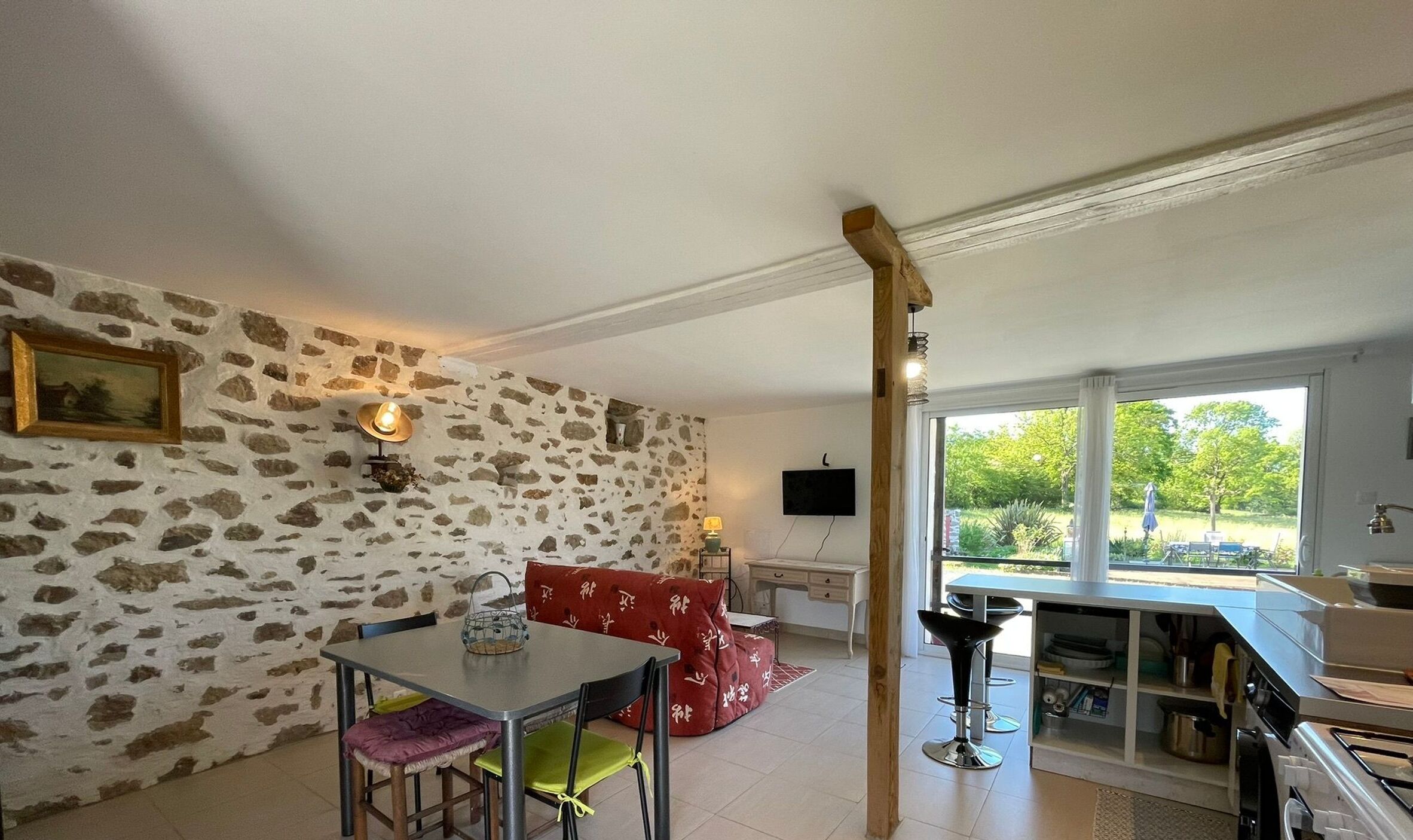 Une pièce avec un mur en pierre, une table à manger avec des chaises, un canapé rouge, un téléviseur et une fenêtre donnant sur un paysage verdoyant.