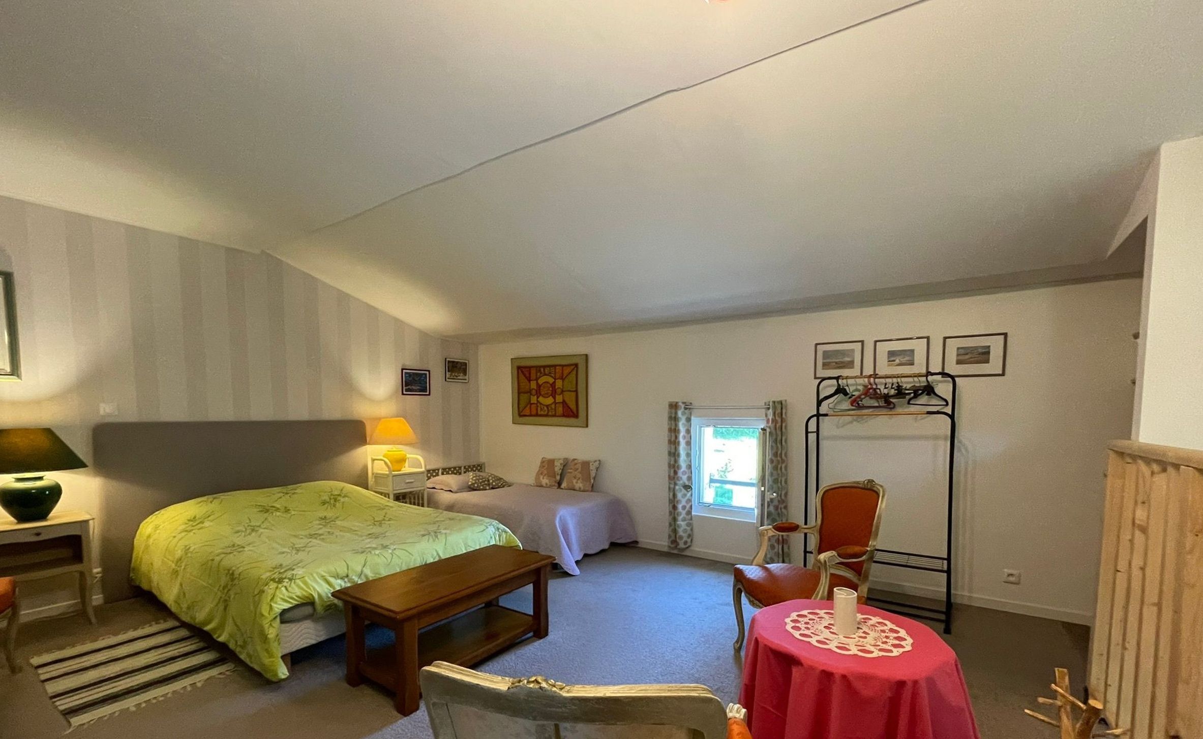 Una camera da letto con un letto, un comodino, una sedia, un tavolino con tovaglia rossa, uno specchio e una finestra con tende.
