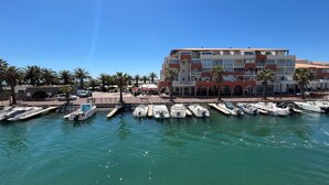 A marina with several boats docked, a building with balconies, and palm trees in the background.