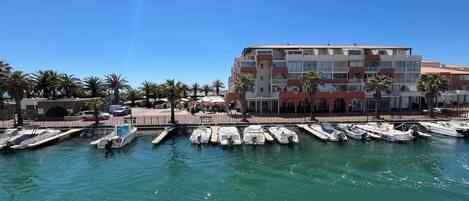 A marina with several boats docked, a building with balconies, and palm trees in the background.