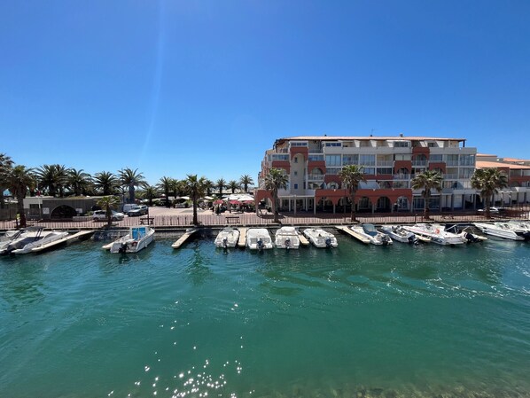 A marina with several boats docked, a building with balconies, and palm trees in the background.
