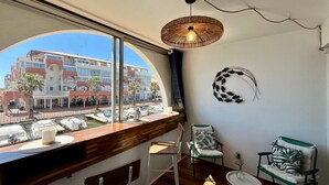 A room with a large window overlooking a marina, a wooden bar area, two chairs, a round table, and a wall-mounted light fixture.