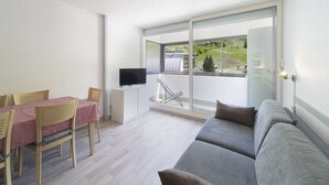 Un salon moderne avec un canapé gris, une table à manger et des chaises, un téléviseur à écran plat et un balcon offrant une vue sur un paysage verdoyant.