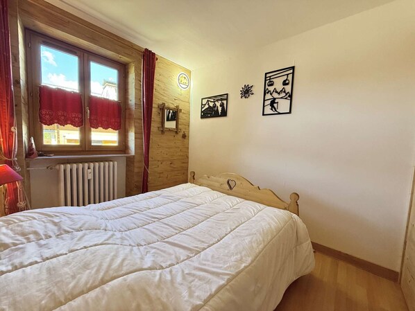 A bedroom with a wooden headboard, a bed with white bedding, a window with red curtains, a wall clock, and two framed pictures.