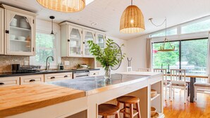 Une cuisine moderne avec des meubles blancs, un grand îlot central doté de tabourets et de grandes fenêtres offrant une vue sur la verdure environnante.