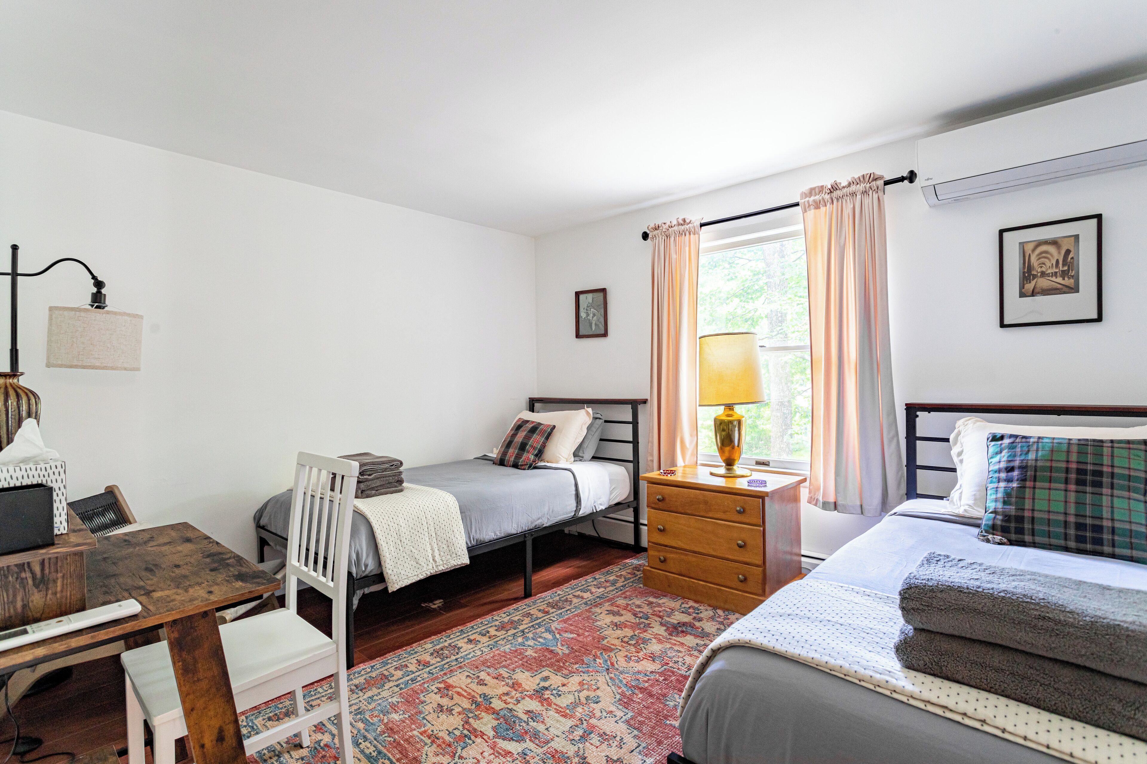 A bedroom with two single beds, a wooden desk, a chair, a small table, a lamp, and a window with curtains.