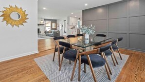 Een moderne eetruimte met een glazen tafel, omringd door donkere fauteuils, een decoratieve zonnespiegel aan de muur en een grijs visgraat tapijt.