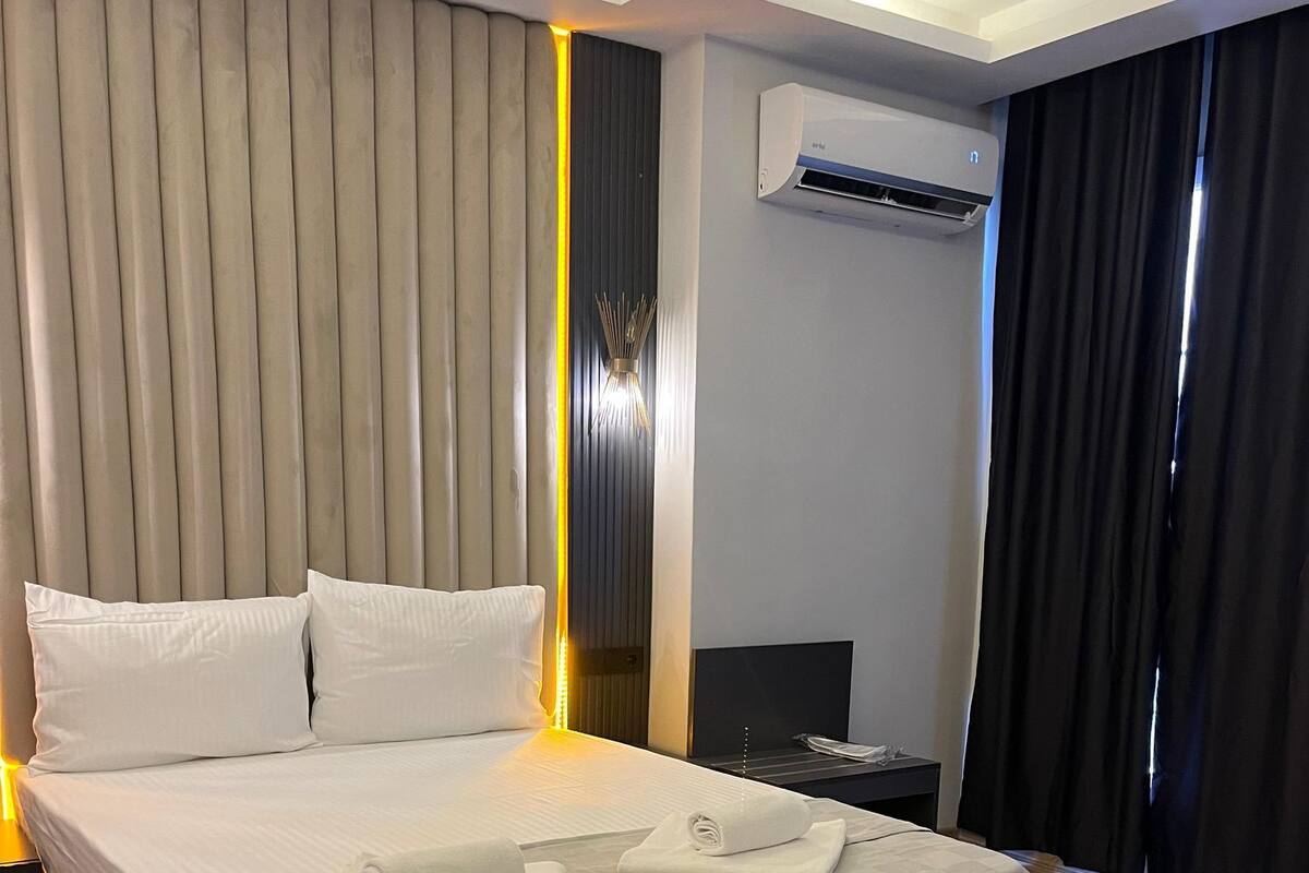 A neatly made bed with white linens and towels, a modern air conditioning unit, and a wooden floor.