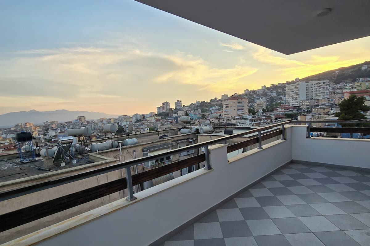 A balcony with a checkered floor, overlooking a cityscape with buildings and a distant mountain range.