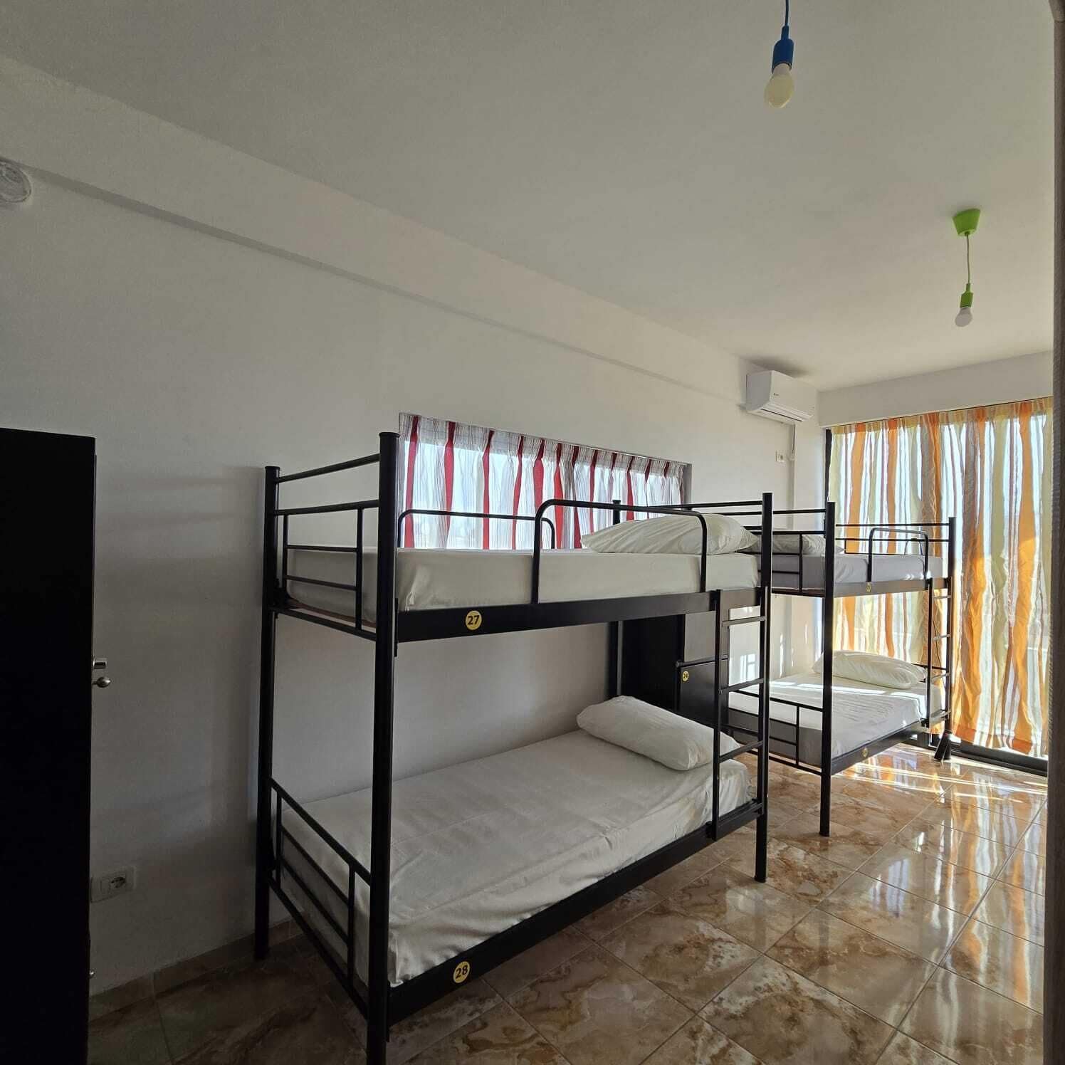Habitación con literas, una puerta, una ventana con cortinas y suelo de baldosas.