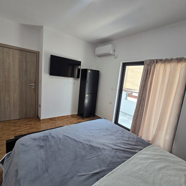 A room with a bed, a television, a refrigerator, and a sliding glass door to a balcony. - Lazy Roo Hostel Saranda (Sarande)