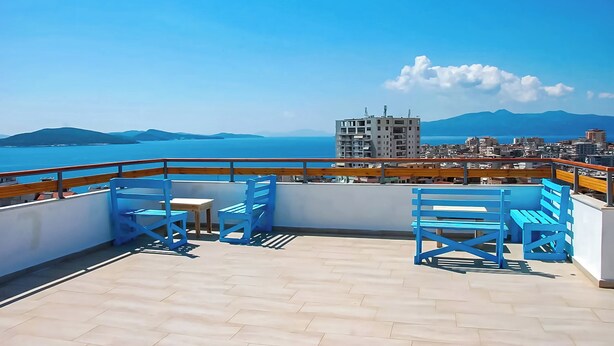 A rooftop terrace with blue chairs and a table, overlooking a coastal cityscape and the sea.