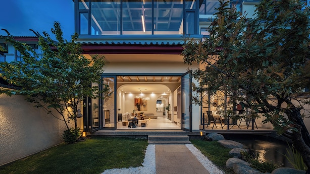 A modern house with a glass facade, a well-maintained garden, and a visible interior with a dining area and kitchen.