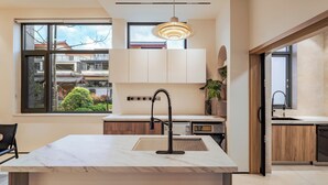 A modern kitchen with a marble countertop, a built-in oven, and a sink. There is a window with a view of a traditional building outside.