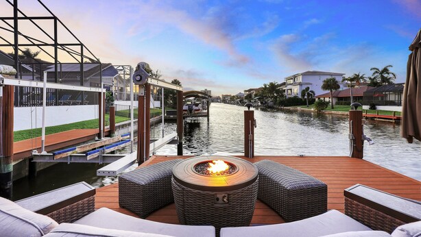 Eine Terrasse mit Feuerstelle, Korbmöbeln und Blick auf einen Kanal mit Gebäuden und Palmen.