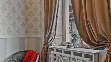 A room with patterned wallpaper, a wicker chair with a red throw, a white console table with decorative items, and brown curtains.
