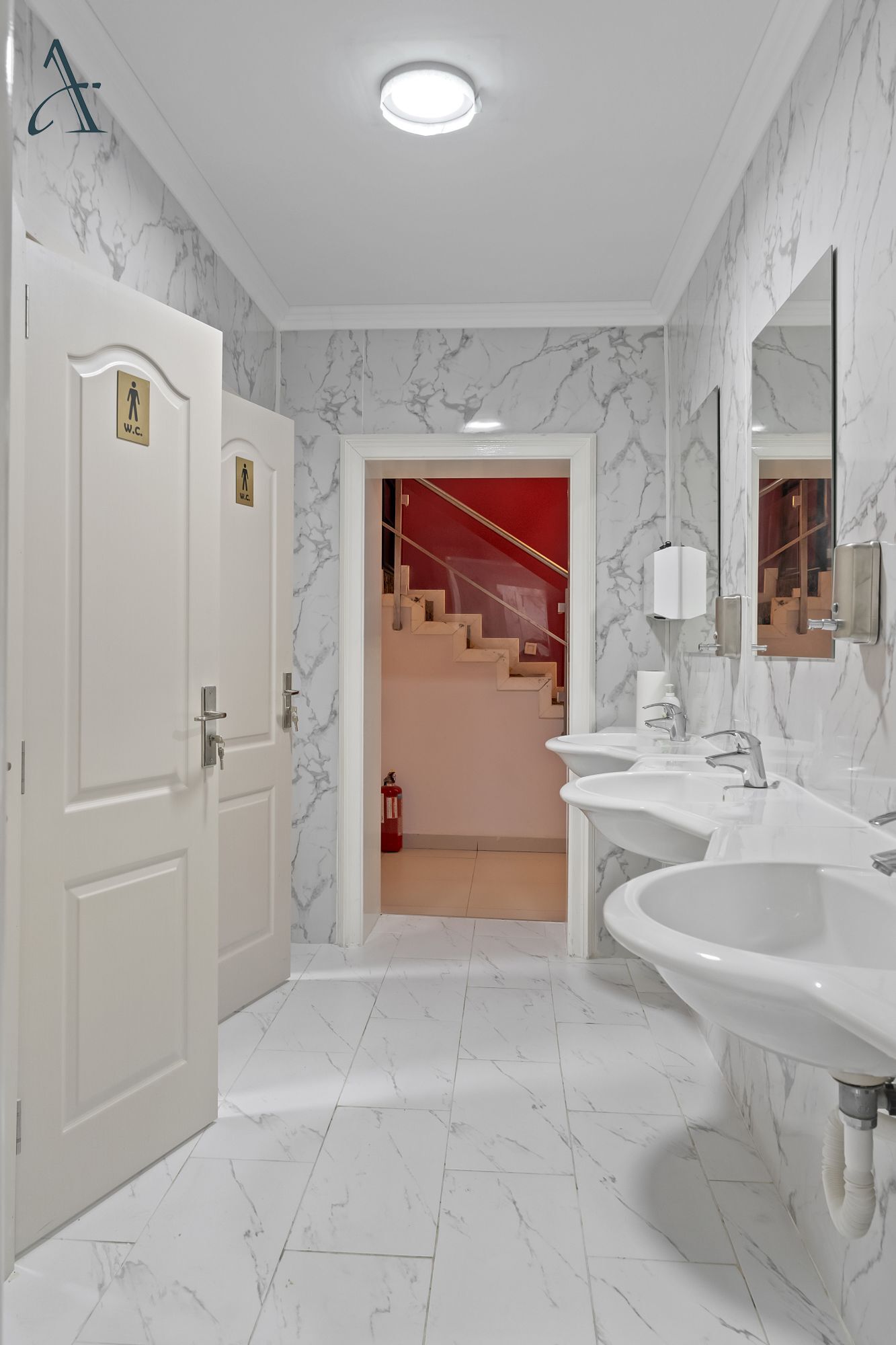 A modern bathroom with white marble walls and floors, two sinks, and a red staircase in the background.
