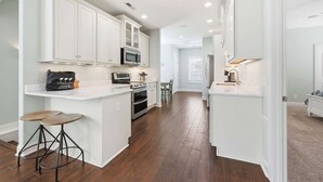 A kitchen with white cabinetry, a microwave, an oven, and a coffee machine. There are two stools and a dining table in the background.