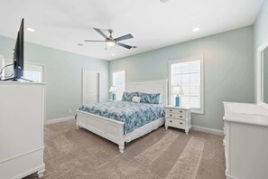 Ein Schlafzimmer mit Bett, Nachttischen, Kommode, Deckenventilator und Fenstern.