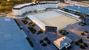 Un grand stade où se pratique divers sports, doté d’un terrain central et entouré d’un immeuble à plusieurs étages, surmonté d’un toit plat.