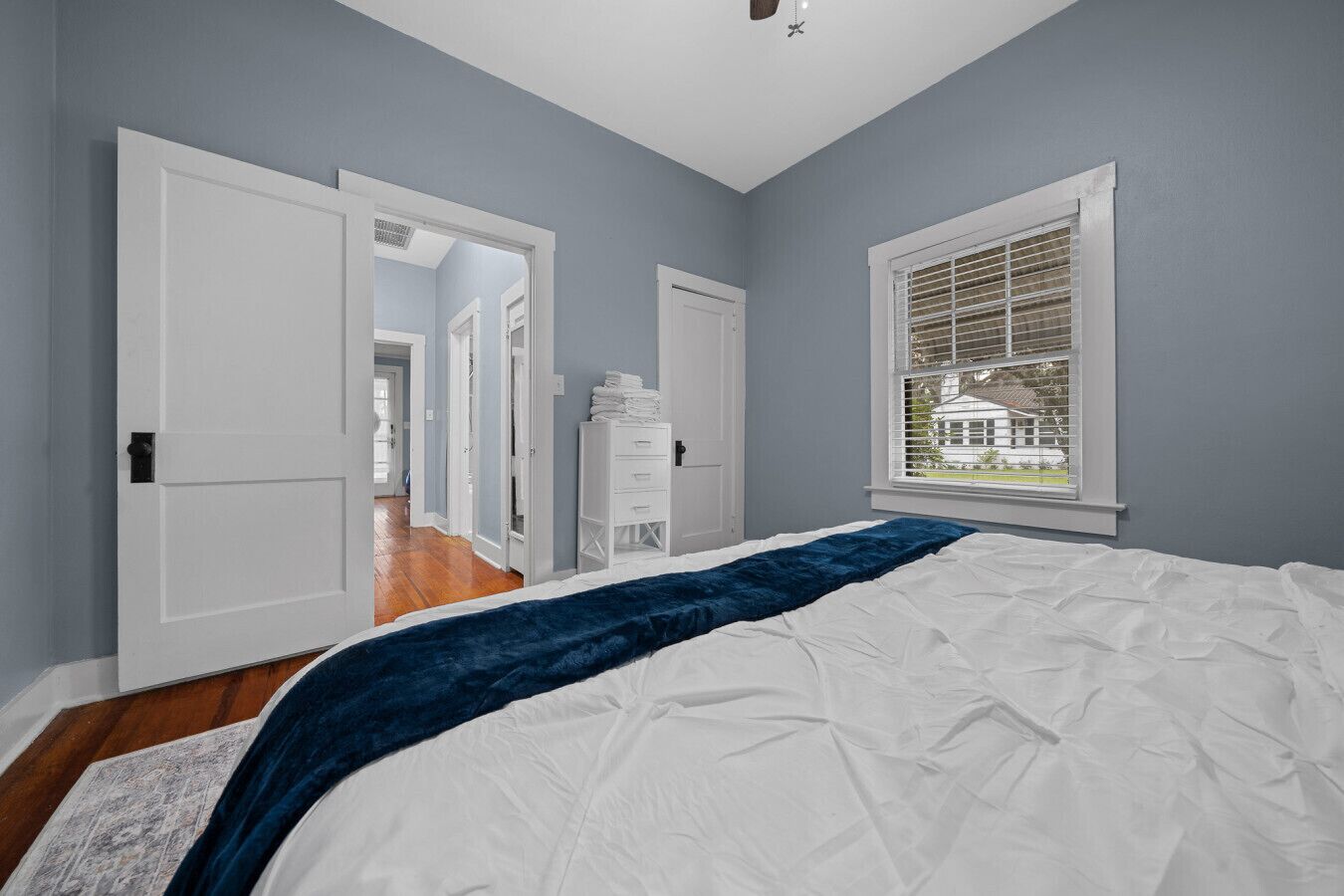 A bedroom with a large bed, a window with blinds, and a view of a house outside.
