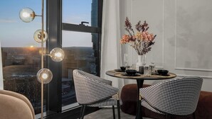 Ein moderner Essbereich mit einem runden Tisch für zwei Personen, einer Blumenvase und Blick auf die Stadt durch große Fenster.