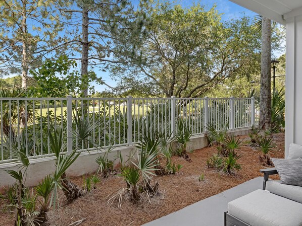 A covered outdoor area with a white fence, palm plants, and a grey sofa.