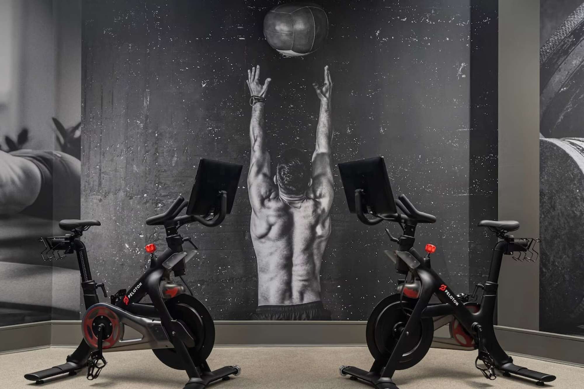 Two stationary bikes in a room with a large wall mural depicting a muscular figure reaching upwards.