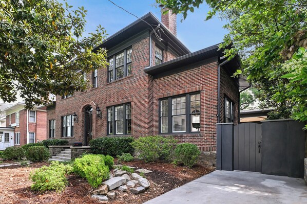 A two-story brick house with a front porch, a gated driveway, and a well-maintained garden with shrubs and rocks.