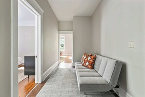 A hallway with a grey sofa, patterned rug, and a door leading to a bedroom.