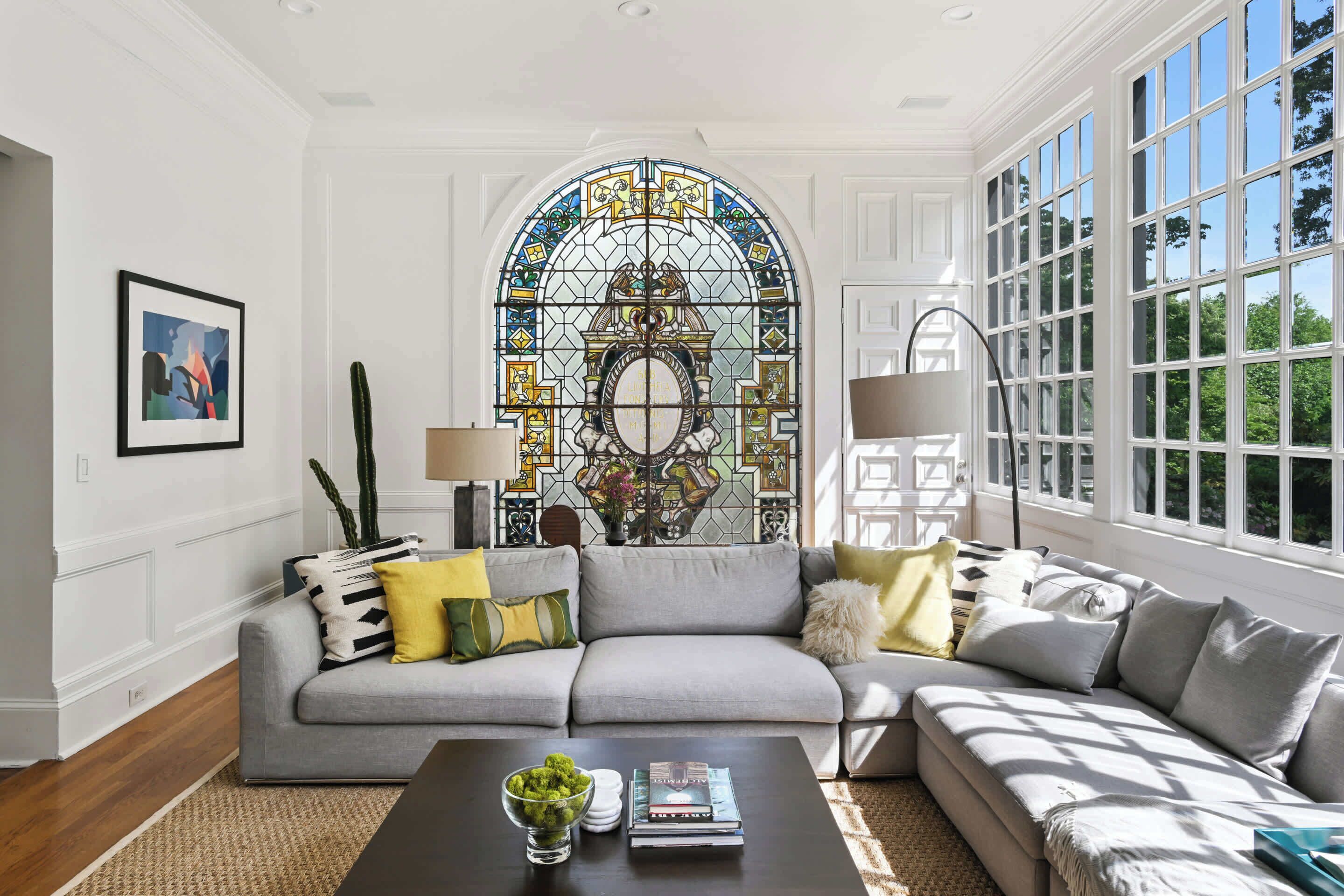 Un salón con un sofá gris, una mesa de centro con libros y una planta, un amplio ventanal de vitral y vistas al verdor exterior.
