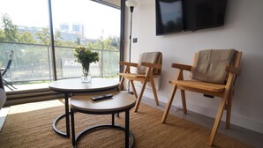 A modern room with a round table, two wooden chairs, a TV mounted on the wall, and a large window with a view of buildings and trees.
