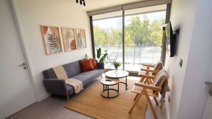 A modern living room with a sofa, coffee table, and chairs, large windows with a view of trees and buildings, and artwork on the wall.