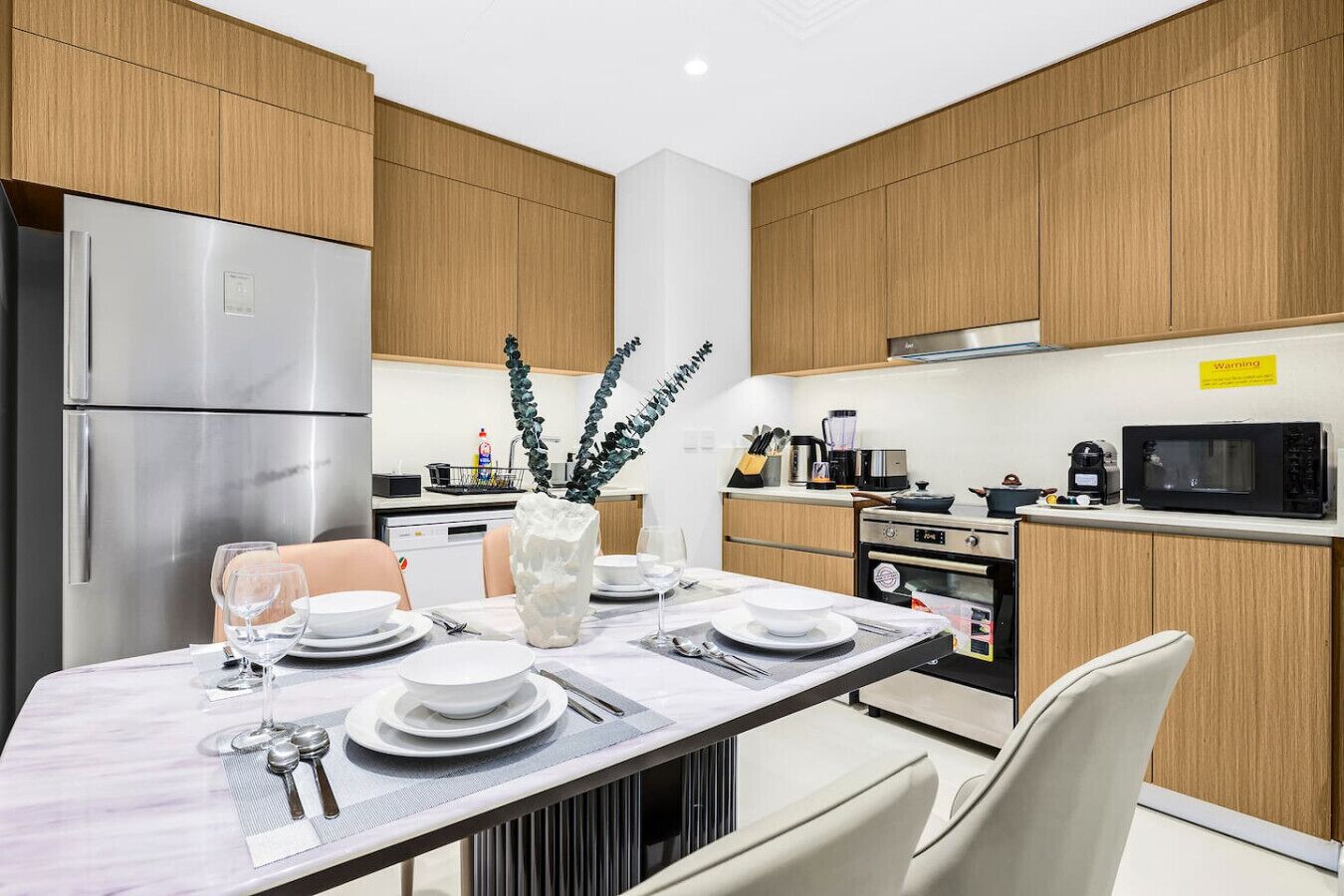 Una cocina moderna con armarios de madera, electrodomésticos de acero inoxidable y una zona de comedor con platos y cubiertos blancos.