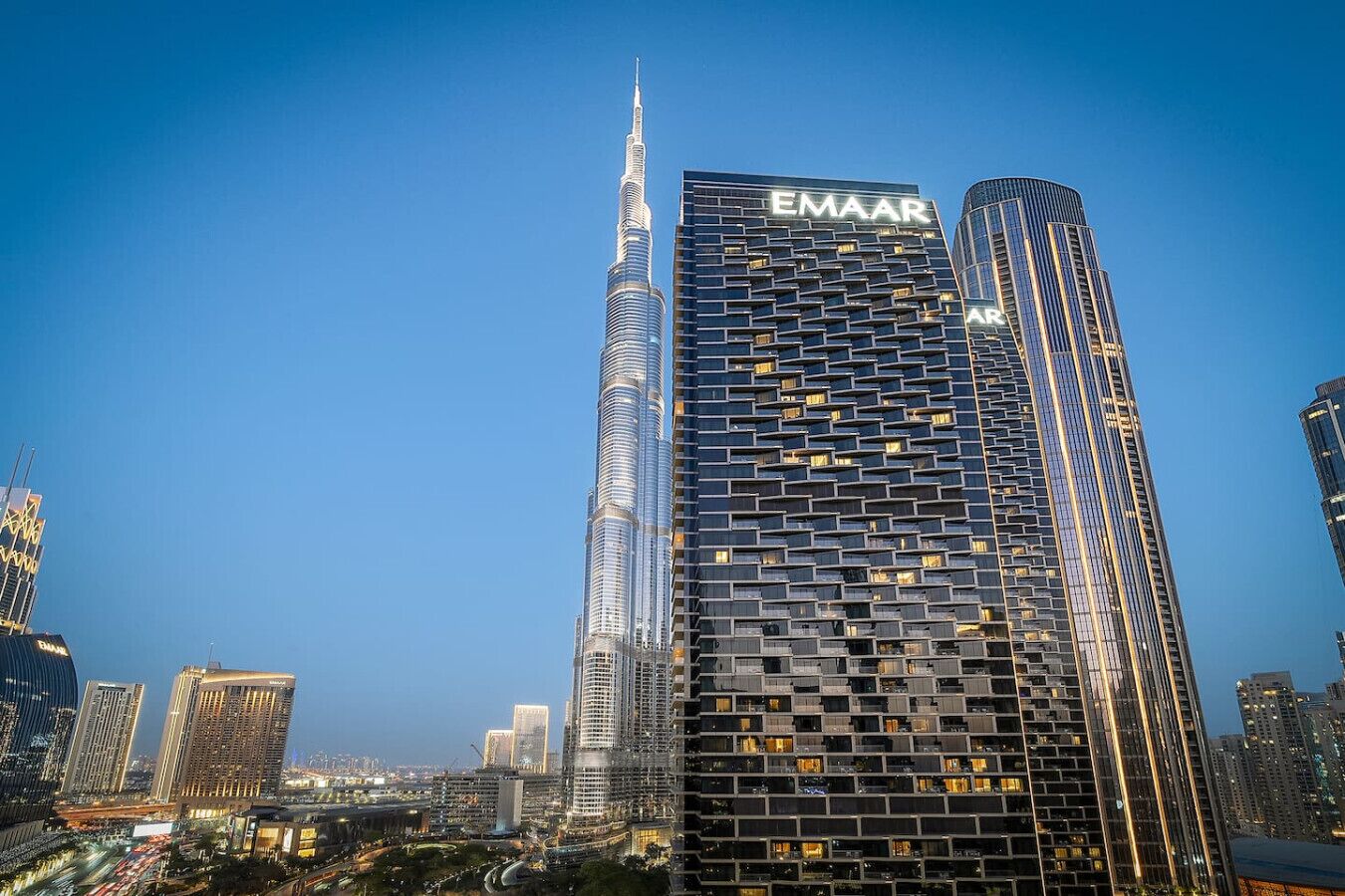 A modern skyscraper with a distinctive spire, adjacent to a building with a grid-like facade and the name 'EMAAR' visible.