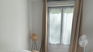 A neatly made bed with a white duvet, beige curtains, and a standing fan. - Charming T2 on the edge of the Seine with terrace (Mantes-la-Jolie)