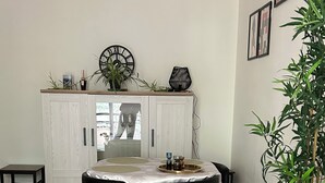 A dining area with a round table, two black chairs, a wooden floor, a white cabinet with a clock, and a potted plant. - Charming T2 on the edge of the Seine with terrace (Mantes-la-Jolie)