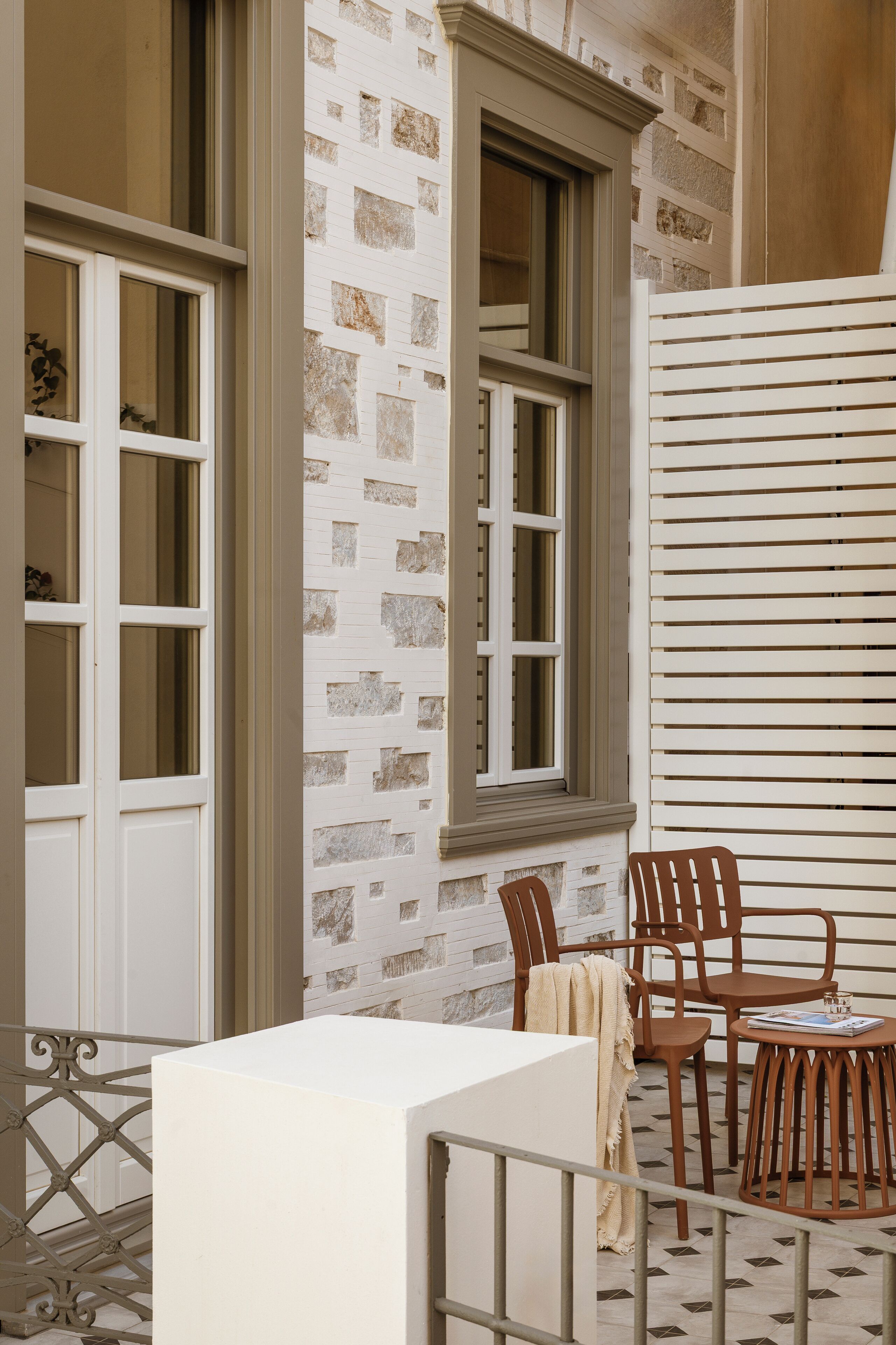 A balcony with a white table and chairs, a window with a view of the building's facade, and a patterned floor.