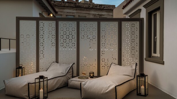 A modern bedroom with a sectional sofa, two bedside tables with candles, and a decorative screen. - Alma Syros (Syros)
