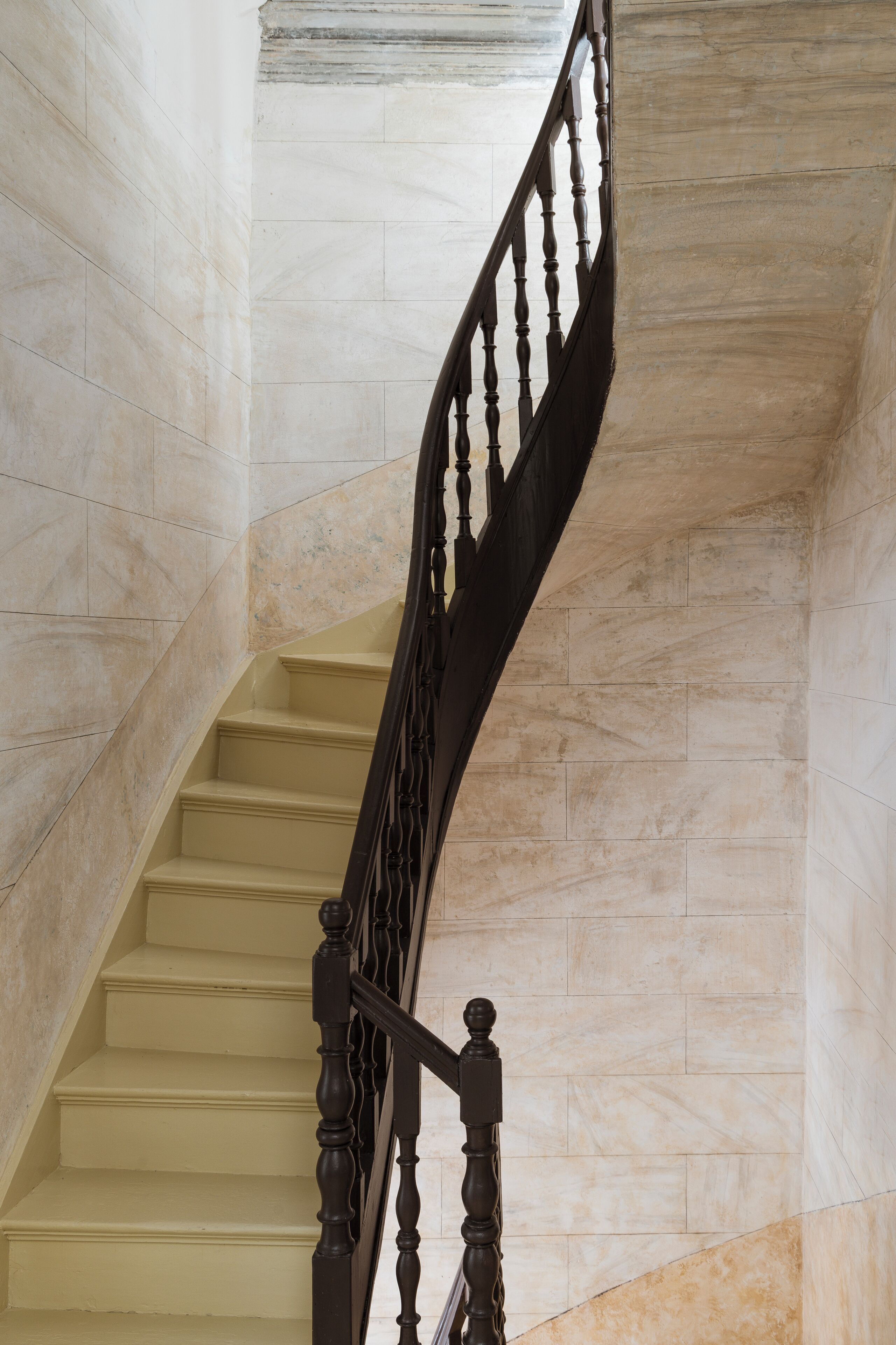A spiral staircase with beige steps and dark wooden balusters, leading upwards in a building with light-colored stone walls.