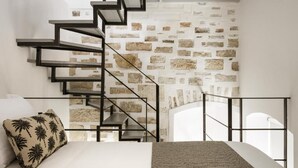 A bedroom with a bed, a pillow, a textured rug, a staircase with a stone wall, and a window. - Alma Syros (Syros)
