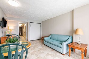 A living room with a sofa, a dining table with chairs, a TV mounted on the wall, and a door leading to another room.