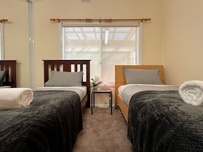 A room with two beds, a small table with a lamp, and a window with blinds.