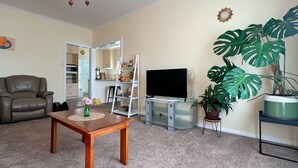A living room with a brown leather chair, a wooden coffee table, a TV on a stand, a white ladder shelf, and large green plants. - Coleman Comfort Hub  - close to City&Air port (Moonah)