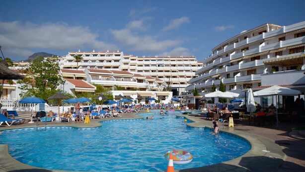 Una grande piscina circondata da lettini e ombrelloni, con un edificio di un hotel sullo sfondo.