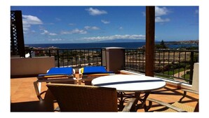 A balcony with a table and chairs, overlooking a coastal view with buildings and a clear sky.