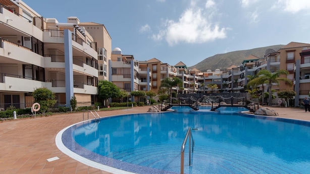 A swimming pool surrounded by multi-story buildings with balconies, a bridge over the pool, and a mountainous backdrop.