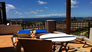 A balcony with a table and chairs, overlooking a coastal view with buildings and a clear sky.